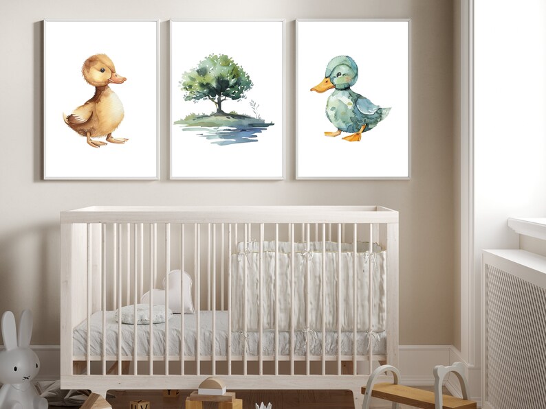 May include: Three watercolor prints featuring a brown duck, a blue duck, and a green tree with a blue pond. The prints are framed in white and hung on a white wall above a white crib.