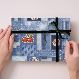 Op de afbeelding: Een rechthoekige geschenkdoos verpakt in denim-stijl papier met patches, kersen en bloemmotieven. Een zwart fluwelen lint en een donkergroene strik beveiligen het pakket. De doos wordt door twee handen vastgehouden.
