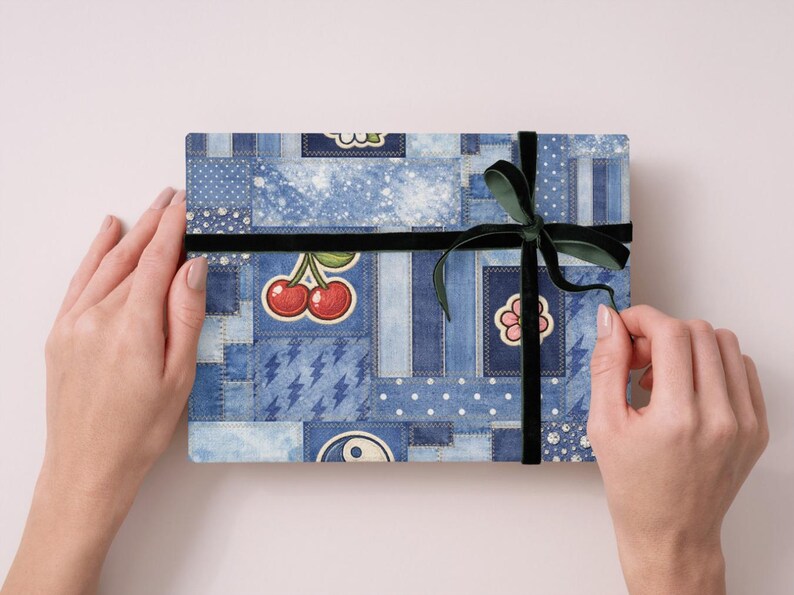 Op de afbeelding: Een rechthoekige geschenkdoos verpakt in denim-stijl papier met patches, kersen en bloemmotieven. Een zwart fluwelen lint en een donkergroene strik beveiligen het pakket. De doos wordt door twee handen vastgehouden.