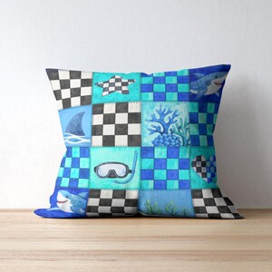 May include: Square throw pillow with a patchwork design featuring sharks, a diving mask, coral, and checkered patterns in shades of blue, teal, and black. The pillow is placed on a wooden surface.