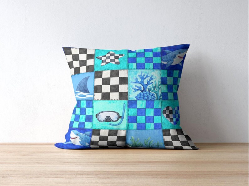 May include: Square throw pillow with a patchwork design featuring sharks, a diving mask, coral, and checkered patterns in shades of blue, teal, and black. The pillow is placed on a wooden surface.