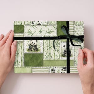 May include: A gift box wrapped in green panda-themed paper with a black velvet ribbon. The paper features panda illustrations, bamboo, and a patchwork design. The box is held by two hands.