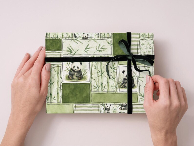 May include: A gift box wrapped in green panda-themed paper with a black velvet ribbon. The paper features panda illustrations, bamboo, and a patchwork design. The box is held by two hands.