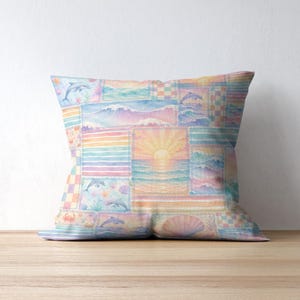 Pode incluir: Almofada decorativa quadrada com um design de patchwork com cenas oce&acirc;nicas. A almofada tem um estilo aquarela com cores pastel, incluindo golfinhos, ondas, pores do sol e conchas. A almofada est&aacute; colocada sobre uma superf&iacute;cie de madeira.