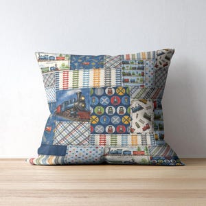 K&ouml;nnte beinhalten: Quadratisches Zierkissen mit einem Patchwork-Design mit bunten Zugillustrationen. Das Kissen hat einen blauen Rand und liegt auf einer Holzoberfl&auml;che. Der Patchwork-Stoff zeigt Bahn&uuml;bergangsschilder und Bahngleise.