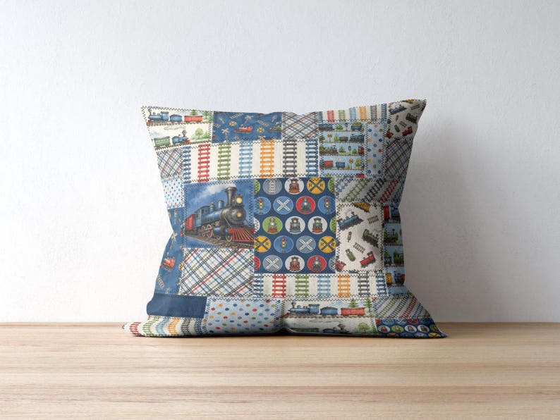K&ouml;nnte beinhalten: Quadratisches Zierkissen mit einem Patchwork-Design mit bunten Zugillustrationen. Das Kissen hat einen blauen Rand und liegt auf einer Holzoberfl&auml;che. Der Patchwork-Stoff zeigt Bahn&uuml;bergangsschilder und Bahngleise.