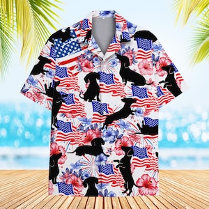 Op de afbeelding: Wit overhemd met knoopsluiting en een patriottisch ontwerp met Amerikaanse vlaggen, vuurwerk en zwarte teckel silhouetten. Rode, witte en blauwe bloemenaccenten maken de feestelijke look compleet. Het shirt heeft korte mouwen en een klassieke kraag.