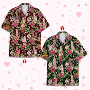 May include: Two short-sleeved button-down shirts with a repeating pattern of phallic figures, flowers, and leaves. One shirt is burgundy, the other is dark green. Both shirts have a white collar and are set against a pink heart background.