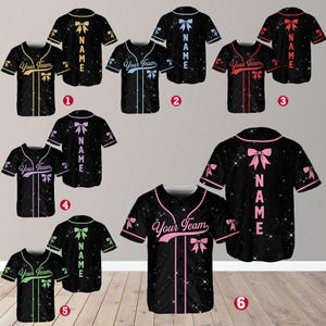 Peut inclure: Six maillots de baseball différents avec un fond scintillant et un nœud rose au dos. Les maillots sont de différentes couleurs, notamment noir, bleu, rouge, vert et jaune. Le texte "Your Team" et "NAME" est imprimé sur les maillots.