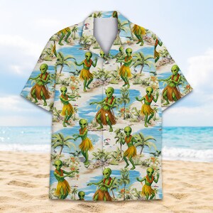 May include: Short-sleeved button-down shirt with a tropical beach scene featuring green aliens in grass skirts. The shirt has a white collar and a repeating pattern of aliens, palm trees, and ocean waves. The background is a light blue sky.