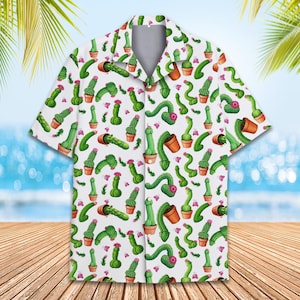 May include: White button-down shirt with a repeating pattern of green cactus illustrations, some potted, some with pink flowers. The shirt has short sleeves and a classic collar, suitable for casual wear. The background shows a beach scene.