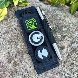 May include: A black fabric pouch with three patches and a silver pen. The patches are a green and black logo, a white circle with a black "G" and the text "EDC", and a white circle with a black "K" shape.