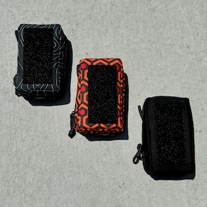 May include: Three small zippered pouches with black velcro patches. The pouches are made of different fabrics: one is black with a wavy pattern, one is orange with a geometric pattern, and one is black.