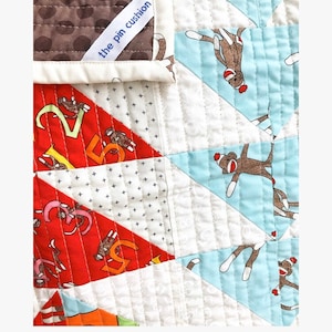 May include: A white fabric quilt with a label that reads "the pin cushion". The quilt is made of different fabrics with various patterns, including red triangles with white numbers and blue triangles with brown monkeys wearing red hats.