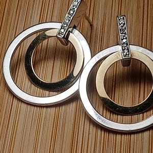 May include: A pair of silver and gold-toned hoop earrings. Each earring features two interlocking circles, one silver and one gold. A rectangular bar, encrusted with small, clear stones, connects the hoops to the earring post.