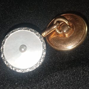 May include: A pair of cufflinks with a gold-tone backing and a white, round face. The face has a pearl in the center and is surrounded by a silver-tone, textured border. The cufflinks are connected by a gold-tone bar.