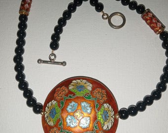 Puede incluir: Collar de cuentas negras con un colgante redondo naranja con un diseño floral en azul, verde y blanco. El collar tiene un cierre de palanca y cuentas cilíndricas decorativas.