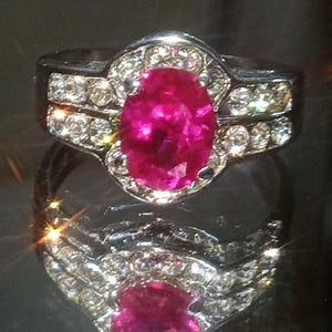 Puede incluir: Un anillo plateado con una gran gema rosa brillante de forma ovalada en el centro, rodeada de piedras más pequeñas y transparentes. Piedras transparentes adicionales están engastadas a lo largo de la banda. El anillo se muestra sobre una superficie reflectante.