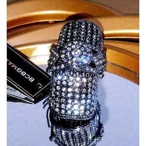 May include: A sparkling, ornate ring with a dark metallic finish, encrusted with numerous clear crystals. The ring features intricate detailing and a bold, eye-catching design. The brand tag is visible in the image.