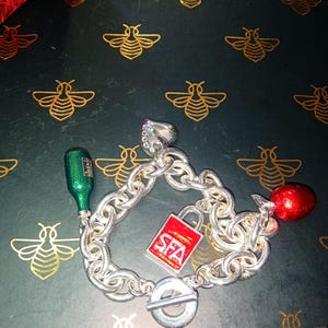 May include: A silver charm bracelet with a toggle clasp. Charms include a green bottle, a red padlock with "SFA" on it, and a red charm. The bracelet is on a dark surface with gold bee designs.