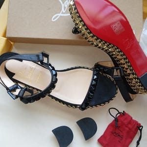 May include: Black sandals with gold and black embellishments, a red sole, and a small red drawstring bag. The sandals have a strap around the ankle and are displayed with two heel protectors.