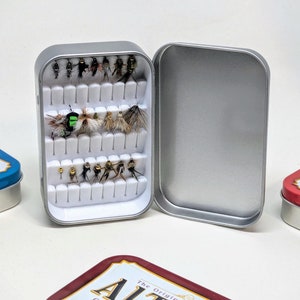 May include: A silver metal fly fishing box with a white foam insert containing a variety of flies. The box is open and the lid is laying on the table. The box is sitting on a white surface.