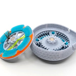 May include: A light gray and blue fly fishing reel with a gear mechanism. The reel is open, revealing the internal gears. The spool is blue and orange, with a fly attached. The reel is designed for fly fishing.