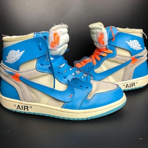 K&ouml;nnte beinhalten: Paar High-Top-Sneaker in Blau, Creme und Grau mit orangefarbenen Akzenten. Das Wort "AIR" in Schwarz ist auf der Zwischensohle zu sehen. Der Nike-Swoosh ist auf der Seite sichtbar. Die Zunge hat ein Nike Air-Etikett. Die Schuhe haben eine cremefarbene Sohle.