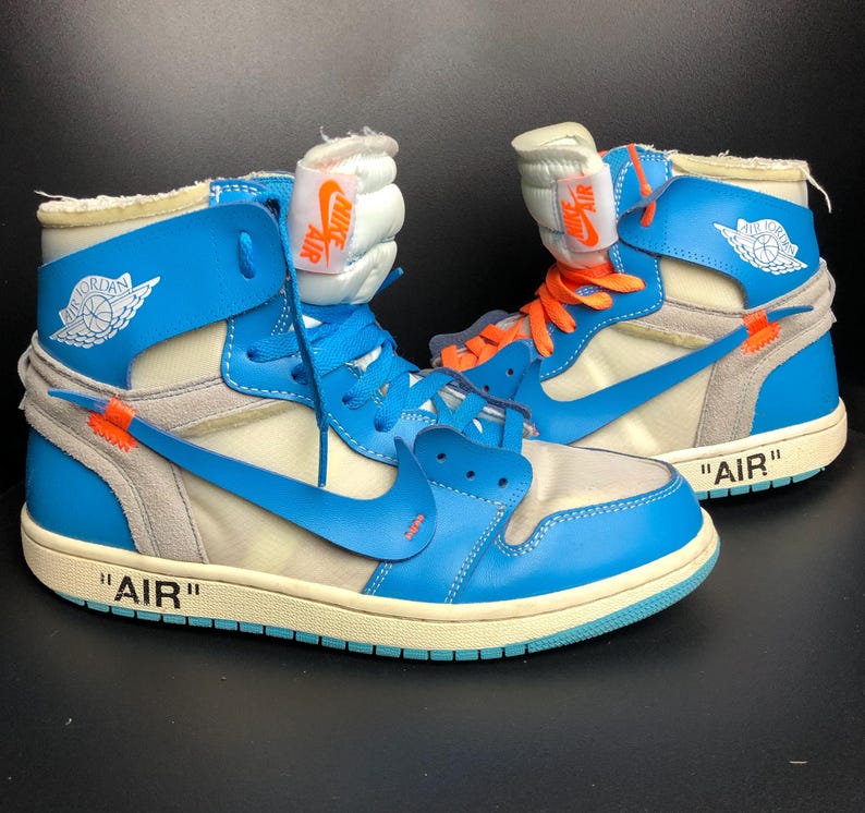 K&ouml;nnte beinhalten: Paar High-Top-Sneaker in Blau, Creme und Grau mit orangefarbenen Akzenten. Das Wort "AIR" in Schwarz ist auf der Zwischensohle zu sehen. Der Nike-Swoosh ist auf der Seite sichtbar. Die Zunge hat ein Nike Air-Etikett. Die Schuhe haben eine cremefarbene Sohle.