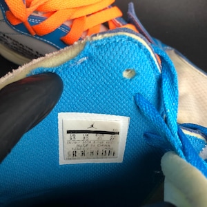 K&ouml;nnte beinhalten: Nahaufnahme eines blau-wei&szlig;en Sneakers mit orangefarbenen Schn&uuml;rsenkeln. Das Innere des Schuhs ist hellblau, mit einem wei&szlig;en Etikett mit Markenname und Gr&ouml;&szlig;enangaben. Der Schuh wird in China hergestellt.