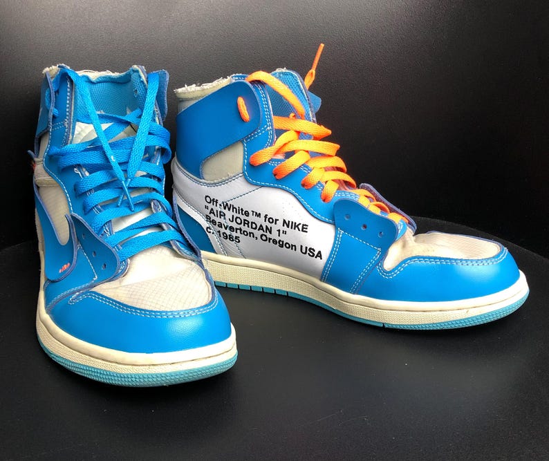 K&ouml;nnte beinhalten: Paar hochgeschnittene Sneaker in Blau, Wei&szlig; und Creme. Die Sneaker haben blaue und orangefarbene Schn&uuml;rsenkel mit dem Text "Off-White for NIKE AIR JORDAN 1" an der Seite. Die Schuhe haben ein Retro-Design.