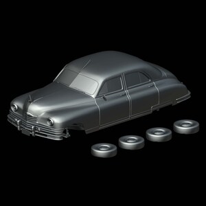 Op de afbeelding: Een grijs automodel in vintage-stijl met vier losse wielen. De auto heeft een klassiek ontwerp met een lange carrosserie, afgeronde spatborden en een prominente grille. Het model staat tegen een zwarte achtergrond.