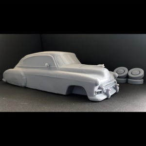 Puede incluir: Un modelo de coche de la década de 1940 impreso en 3D de color gris. El modelo es detallado e incluye un capó, un parabrisas y ruedas.