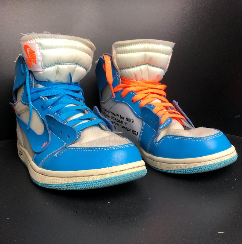 K&ouml;nnte beinhalten: Paar hochgeschnittene Sneaker in Blau, Wei&szlig; und Orange. Die Sneaker haben blaue Schn&uuml;rsenkel, eine wei&szlig;e Zunge und das Nike-Logo. Der Text "OFF-WHITE for NIKE AIR JORDAN 1" ist auf der Seite sichtbar.