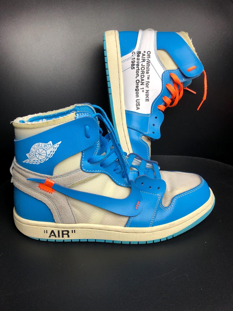 K&ouml;nnte beinhalten: Paar hochgeschnittene Sneaker in Blau, Wei&szlig; und Creme. Die Sneaker haben das Wort "AIR" auf der Zwischensohle, orangefarbene Schn&uuml;rsenkel und den Text "Off-White for NIKE AIR JORDAN 1 Beaverton, Oregon USA C. 1985". Klassisches Design.