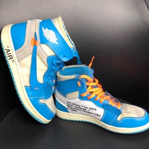 K&ouml;nnte beinhalten: Paar hochgeschnittene Sneaker in Blau, Wei&szlig; und Grau mit orangefarbenen Schn&uuml;rsenkeln. Die Sneaker tragen den Text "AIR" an der Seite und "Off-White for NIKE AIR JORDAN 1". Retro-Design.
