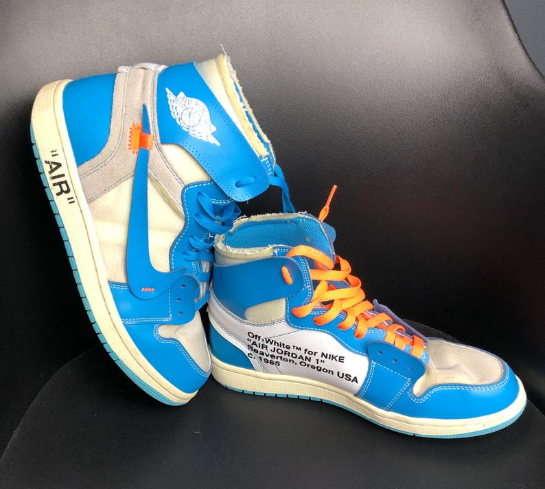 K&ouml;nnte beinhalten: Paar hochgeschnittene Sneaker in Blau, Wei&szlig; und Grau mit orangefarbenen Schn&uuml;rsenkeln. Die Sneaker tragen den Text "AIR" an der Seite und "Off-White for NIKE AIR JORDAN 1". Retro-Design.