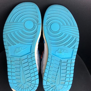 K&ouml;nnte beinhalten: Paar Turnschuhe mit hellblauen Sohlen. Die Sohlen haben ein strukturiertes Muster und das Nike-Logo. Die Oberteile sind wei&szlig; mit blauen und orangefarbenen Akzenten. Die Schuhe sind f&uuml;r sportliche Aktivit&auml;ten konzipiert.
