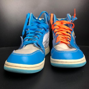 K&ouml;nnte beinhalten: Paar High-Top-Sneaker mit blau-wei&szlig;en Paneelen, mit blauen und orangefarbenen Schn&uuml;rsenkeln. Die Schuhe haben eine hellblaue Sohle und eine wei&szlig;e Mesh-Zunge. Das Design enth&auml;lt Text auf der Seite.