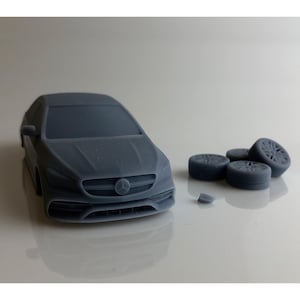 May include: A gray model car with a Mercedes-Benz logo on the front, accompanied by four detached wheels. The car is a detailed miniature, likely 1:24 scale, and is made of a solid, matte material. The wheels have intricate spoke designs.