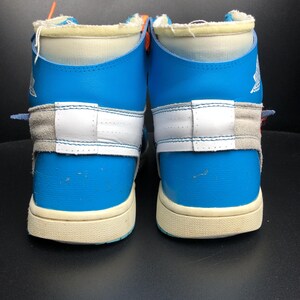 K&ouml;nnte beinhalten: Paar hochgeschnittene Sneaker in Blau, Wei&szlig; und Grau. Die Schuhe haben ein einzigartiges Design mit einer Mischung aus Materialien, darunter Leder und Wildleder. Die Sohlen sind cremefarben und die Schn&uuml;rsenkel sind orange. Sportschuhe.