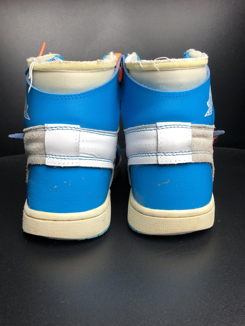 K&ouml;nnte beinhalten: Paar hochgeschnittene Sneaker in Blau, Wei&szlig; und Grau. Die Schuhe haben ein einzigartiges Design mit einer Mischung aus Materialien, darunter Leder und Wildleder. Die Sohlen sind cremefarben und die Schn&uuml;rsenkel sind orange. Sportschuhe.