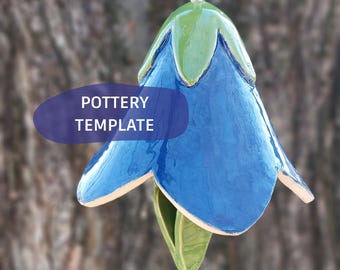 May include: A hanging ceramic bell ornament shaped like a blue flower with green leaves at the top and bottom. The piece features a glossy finish and a string for suspension. A purple graphic overlay on the left side displays the text POTTERY TEMPLATE.