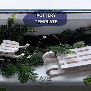 May include: Two small, off-white ceramic sleds with curved runners sit on a windowsill, surrounded by evergreen branches and a string of star-shaped lights. The text "POTTERY TEMPLATE" is in a blue oval.