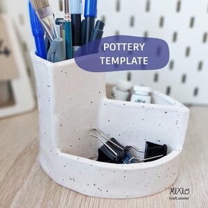 May include: A speckled white pottery organizer with multiple compartments. The organizer holds various art supplies, including paintbrushes, pens, and binder clips. The text "POTTERY TEMPLATE" is displayed on a purple banner. The bottom right corner has the text "XIXIO Craft.atelier".
