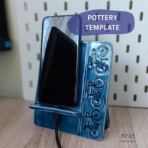 Könnte beinhalten: Türkisblauer Keramik-Telefonständer mit Fahrradmotiven. Er hält ein schwarzes Smartphone, mit einem angeschlossenen Ladekabel. Der Text "POTTERY TEMPLATE" ist sichtbar. Auf einer Holzoberfläche platziert.