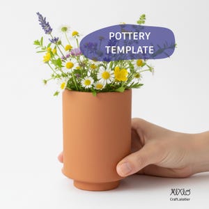 Peut inclure: Un vase cylindrique en terre cuite rempli de fleurs sauvages, dont des marguerites et de la lavande. Le vase a une base arrondie et le texte "POTTERY TEMPLATE" sur un ovale violet. Le vase est tenu par une main.