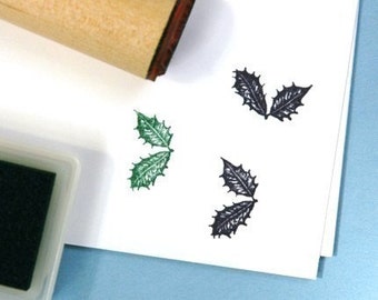 May include: A wooden rolling stamp with a green ink pad and holly leaf stamps in green and black on white paper. The stamps are arranged in a pattern, suggesting a holiday theme.