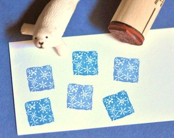 May include: A wooden stamp with a snowflake design is pressed onto a white card, creating blue square patterns. A white seal figurine rests on the card. The stamp handle has the text "RUBBER TAPES MADE IN..." printed on it.