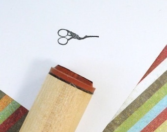 May include: A wooden stamp with a scissors design, next to a stamped image of scissors on white paper. A green and pink polka dot ribbon and a striped paper are in the background. Crafting and stationery supplies.